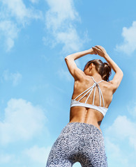 attractive sports woman with tanned fit body posing against blue sky with clouds. Fitness woman in sport wear with muscular back and tight buttocks in leggings. back wiew.