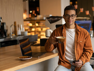 Portrait of stylish African American man freelancer holding cup of coffee and using smartphone and mobile application, looking at camera and smiling, sitting in modern cafe. Coffee break concept