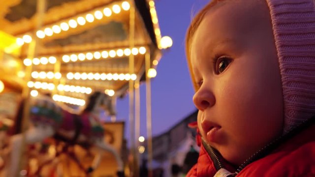 Baby looks at beautiful illuminated carousel in the evening
