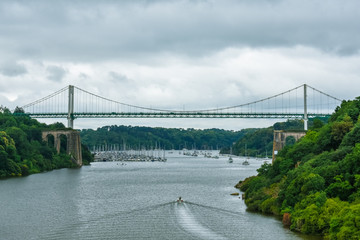 Cable-stayed bridge in a green natural forest, with a dramatic cloudy sky. Motorboat sailing on the river