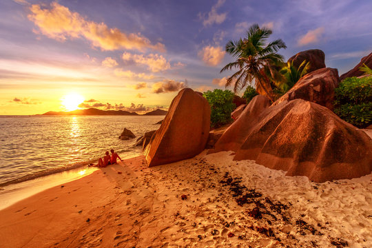 An identified romantic couple on shoreline near shaped granite stones of Anse Source d'Argent with sunset sky. Seychelles honeymoon. Sunlight over the sea on the horizon in La Digue, Seychelles.