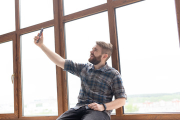 Obraz na płótnie Canvas Side view of a positive handsome hipster guy and taking a selfie on a smartphone while sitting on a windowsill by a large window. Concept of gadgets and modern technologies.