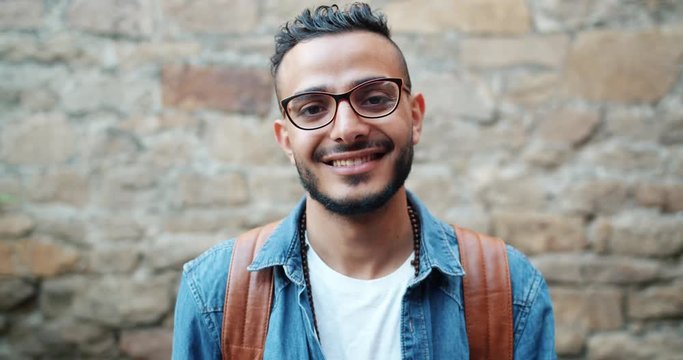 Portrait of attractive young Arab smiling looking at camera standing near brick wall alone wearing stylish clothing, glasses and beads. People and happiness concept.