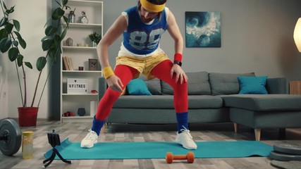 Funny-looking retro fitness instructor shooting exercise videos for vlog at home. Concentrated young man stretching legs and warming up before training.