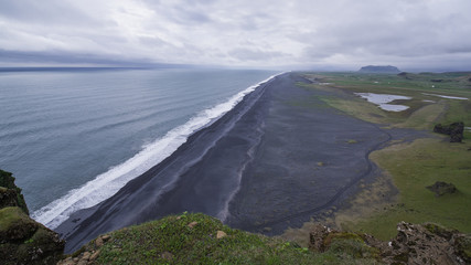 Iceland's Black Beach at Dyrholaey seen from high above. Surf line diagonal. Atlantic on left, black sand beach and grassy lowland right. An inselberg in distance.