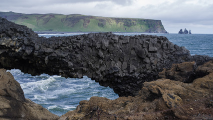 Summer on Iceland at Dyrholaey nature reserve -a look across a bay with a natural arch made by an eroded volcanic rock.  Ocean waves in centre,  mountains above coast with more sharp cliffs in back