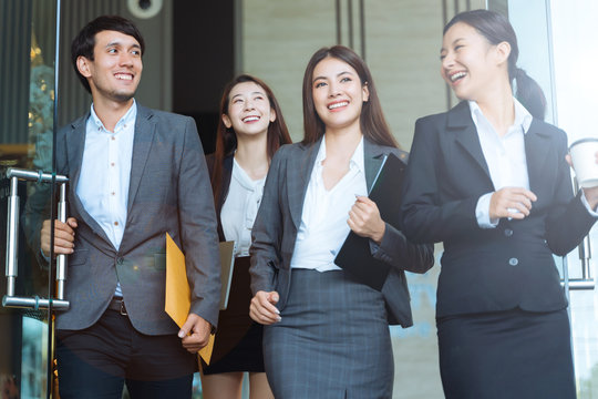 Asian Business People Walking To Go Out Office Building. Young Businessman And Businesswoman Talking And Open The Office Door.