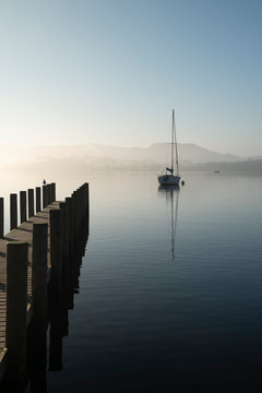 Stunning unplugged fine art landscape image of sailing yacht sitting still in calm lake water in Lake District during peaceful misty Autumn Fall sunrise