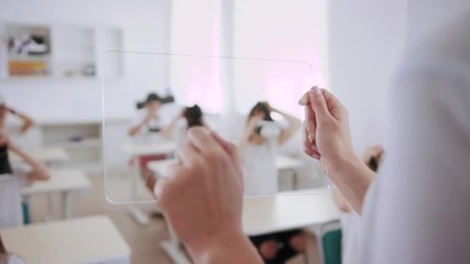 Smart teacher using a transparent tablet augmented reality in the classroom. Close-up of a young woman pressing on a transparent virtual screen for students to wear VR headsets for a futuristic lesson - Powered by Adobe