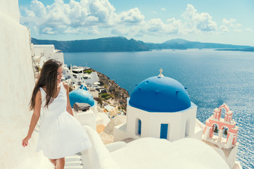 Santorini travel tourist woman on vacation in Oia walking on stairs. Asian girl in white dress visiting the famous white village with the mediterranean sea and blue domes. Europe summer destination.