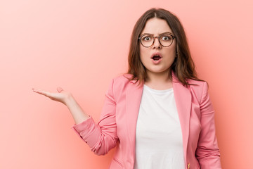 Young plus size business caucasian woman impressed holding copy space on palm.