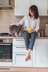 woman sitting on kitchen table with cat drinking tea from yellow mug