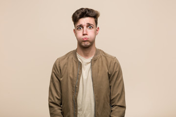 Young caucasian man wearing a brown jacket blows cheeks, has tired expression. Facial expression concept.