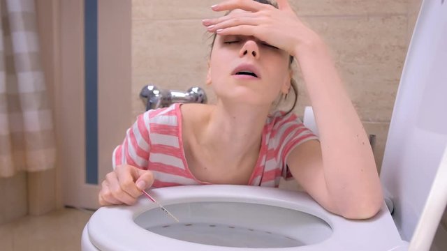 Pregnant young tired woman with pregnancy test in hand is vomiting in toilet sitting on the floor at home, front view.