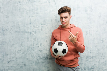 Young sporty man holding a soccer ball pointing with finger at you as if inviting come closer.