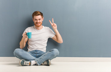 Young redhead student man sitting on the floor fun and happy doing a gesture of victory. He is holding a coffee mug.