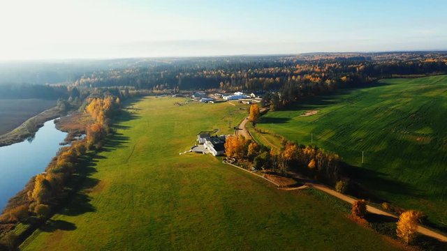Drone flying above beautiful autumn sunrise field, lake and forest revealing big farm ranch and mansion on sunny day.