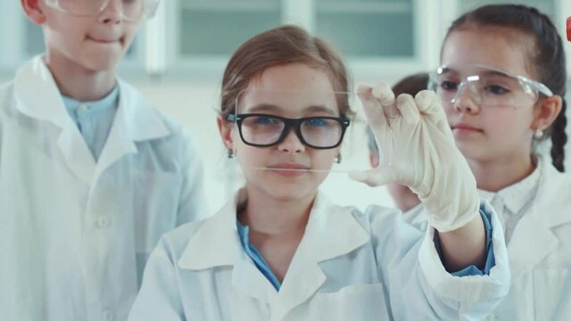 Cute smart girl in glasses holding a virtual invisible smartphone in her hand in the science classroom. Portrait of excited young kids scientists developing a futuristic gadget in a lab.