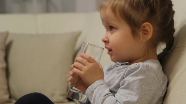 Little child drinking water from a glass sitting on a sofa in the room