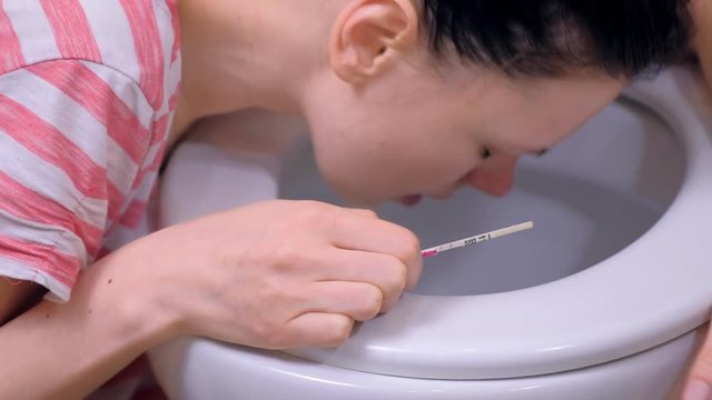 Morning sickness. Young pregnant tired woman with pregnancy test in hand is vomiting in toilet at home, closeup side view.