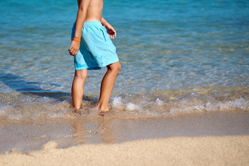 Cute European boy in a blue swimming shorts is standing in  the sea. He is enjoying his summer holidays. Back view.