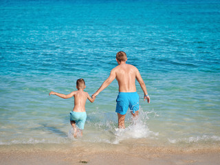 Dad and son are running in to the ocean, they are happy during their summer vacations. - 275983663