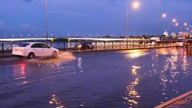 Car traffic with big splashes on the flooded city street. Large puddle on the asphalt road at night. Road after the rain. Emergency - water drains cannot cope with heavy rain, deluge.