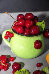 Sweet cherries in a green mug spattered with drops of cool water, next to a gray background a scattering of berries and apricots. 