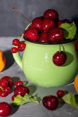 Sweet cherries in a green mug spattered with drops of cool water, next to a gray background a scattering of berries and apricots. 