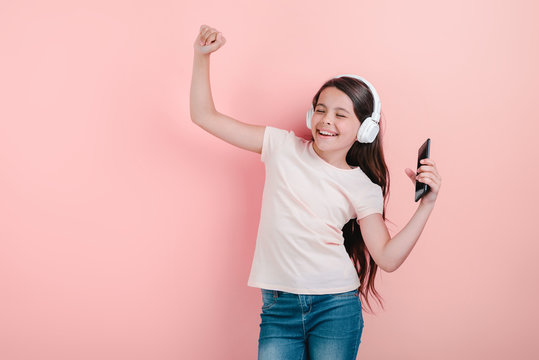 A dancing girl with closed eyes in earphones listening to music and sing with hand up holding telephone in one arm