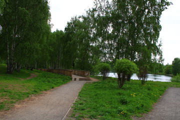 City park, behind the green trees you can see the pond. The alley of the park continues with a wooden bridge.