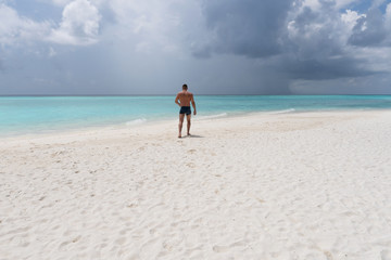 Strong young guy walking on the white sand in the turquoise water.
