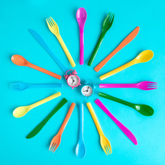 Top view of multicolored plastic spoons, forks and knives with blue and pink alarm clocks isolated on blue.