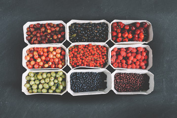 Healthy summer fruit variety. Sweet cherries, strawberries, blackberries, gooseberries, red currant and black currant in paper boxes over a rustic dark stone background. Top view