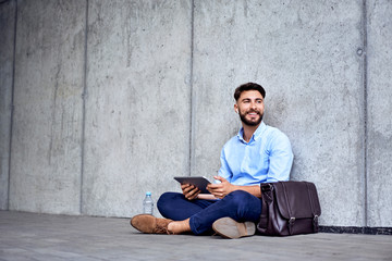 Smilling young businessman in wireless headphones sitting with tablet and looking away