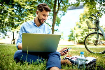 Happy young businessman using laptop and phone while sitting in the park during lunch break