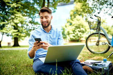 Young entrepreneur sitting in park with laptop and bicycle using smartphone