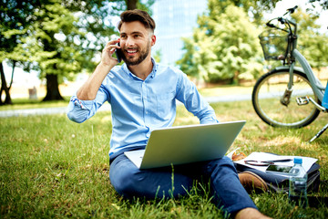 Young businessman talking on phone while sitting in the park with laptop nad bicycle