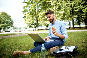 Young businessman having sandwich for lunch in the park and working on laptop