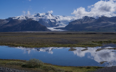 Polar summer in Iceland. Sunlit Mountains covered partly by snow reflect in a lake together with white clouds. A glacier visible across a patch of green between the mountain peaks.