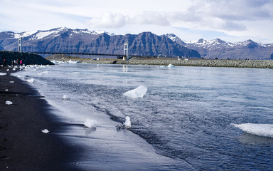 Iceland summer - Jokulsarlon Diamond Beach. Icebergs flowing into Atlantic, some washed up on the black sand beach. Picture taken from the sea towards the lagoon and inland mountains. Bridge and cars 