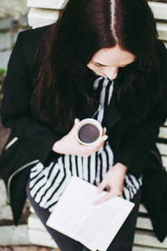 Smiling brunette girl sitting on a bench in a park and reading a soft-cover book.