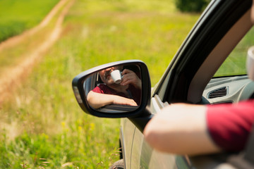 A man sitting in the car and drinking coffee. In the mirror you can see the reflection of his face and a coffee Cup. The background shows a dirt road and a green field.  Copy space. 
