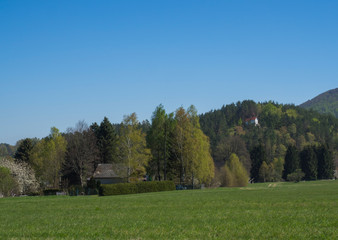Fototapeta na wymiar Spring landscape with view on village Marenice in Lusitian mountains with small chapel on hill, lush green grass meadow, deciduous and spruce tree forest and hills, blue sky background