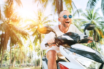 Happy smiling male tourist in sunglasses riding motorbike scooter during his tropical vacation under palm trees with shining sun rays. © Soloviova Liudmyla