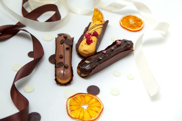 A close-up of a set of three eclairs with different fillings and design isolate on a white surface decorated with chocolate drops, dried oranges and brown silk ribbon
