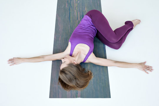 Young woman working out doing yoga exercise on floor variation of Belly Twist Pose, Jathara Parivartanasana. Top view on white floor. Full length