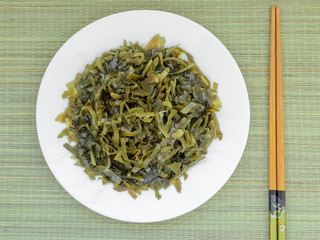 Sliced pickled kelp on a white plate and chopsticks on a green wicker table mat. Edible seaweed contains a lot of iodine and microelements. Seafood, vegetarian nutrition and healthy eating.