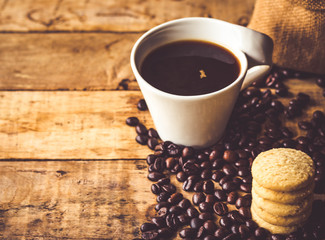 Black coffee In the cup white have roasted coffee beans In a cloth bag sack and dispersedly are on table, have milk cookies, place stacked 5 pieces, all are placed on a wooden table.
