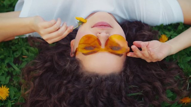 Outdoor flat lay, top view close up portrait of young beautiful happy smiling curly lady wearing stylish orange sunglasses, model lying on the green grass, holding dandelion